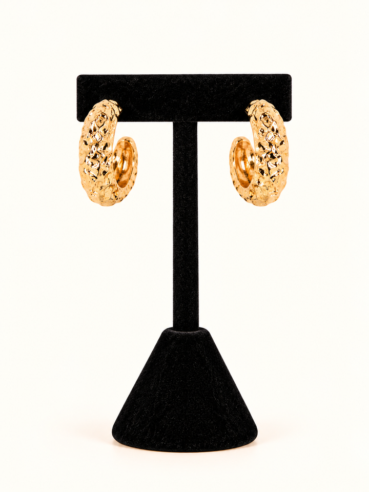 Gold-tone hammered hoop earrings with a bold, textured finish and 1-inch open hoop silhouette displayed on a black velvet T-bar jewelry stand against a cream background.