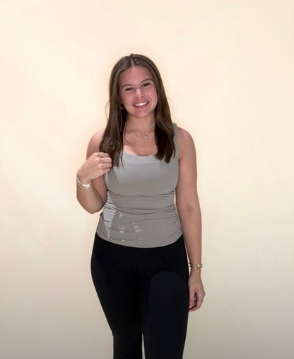 Young woman modeling a taupe fitted tank top with black leggings, posing with a light neutral background.