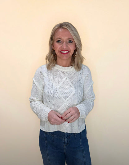 Front view of a woman in a white cable knit sweater and blue jeans, smiling with hands clasped against a cream backdrop.