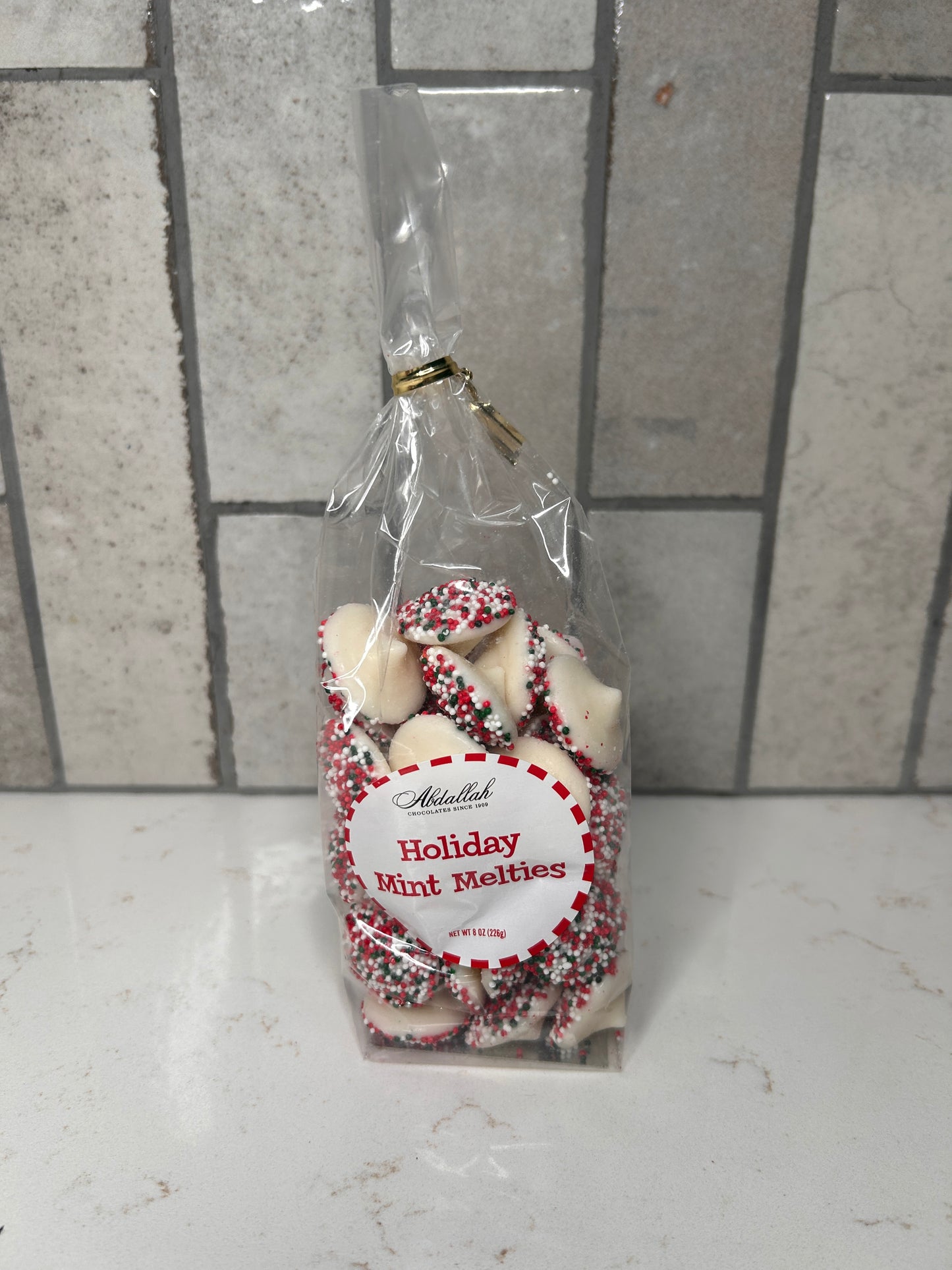 Clear bag of white chocolate holiday mint melties with red, green, and white sprinkles, labeled ‘Holiday Mint Melties,’ displayed on a light countertop with neutral tile backsplash.
