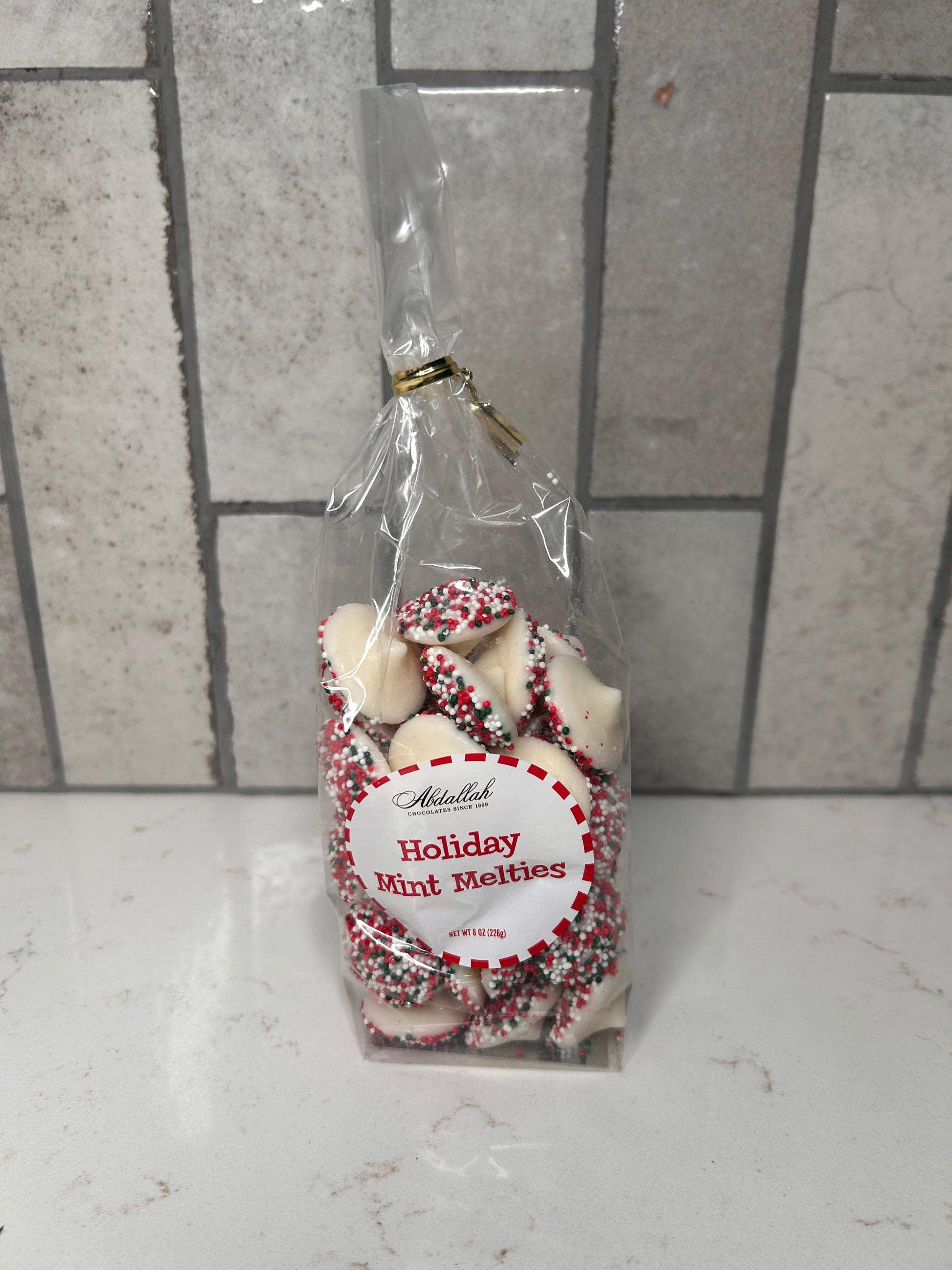 Clear bag of white chocolate holiday mint melties with red, green, and white sprinkles, labeled ‘Holiday Mint Melties,’ displayed on a light countertop with neutral tile backsplash.