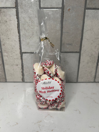 Clear bag of white chocolate holiday mint melties with red, green, and white sprinkles, labeled ‘Holiday Mint Melties,’ displayed on a light countertop with neutral tile backsplash.