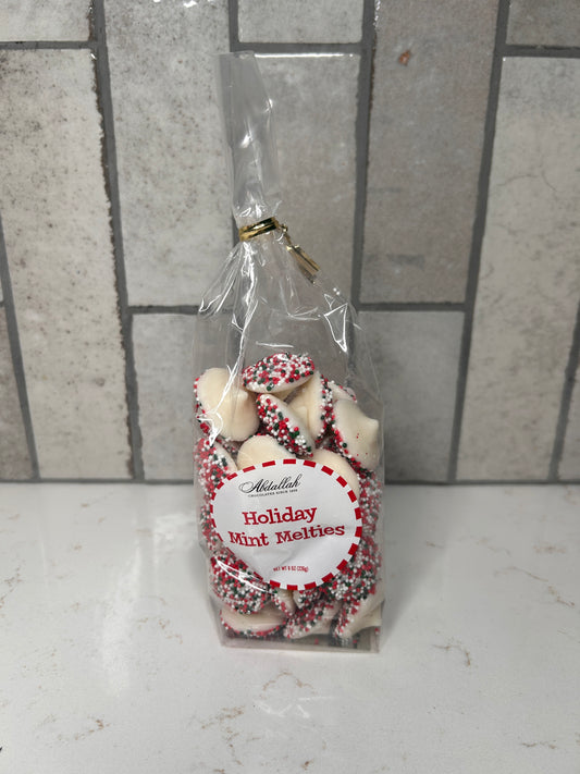 Clear bag of white chocolate holiday mint melties with red, green, and white sprinkles, labeled ‘Holiday Mint Melties,’ displayed on a light countertop with neutral tile backsplash.