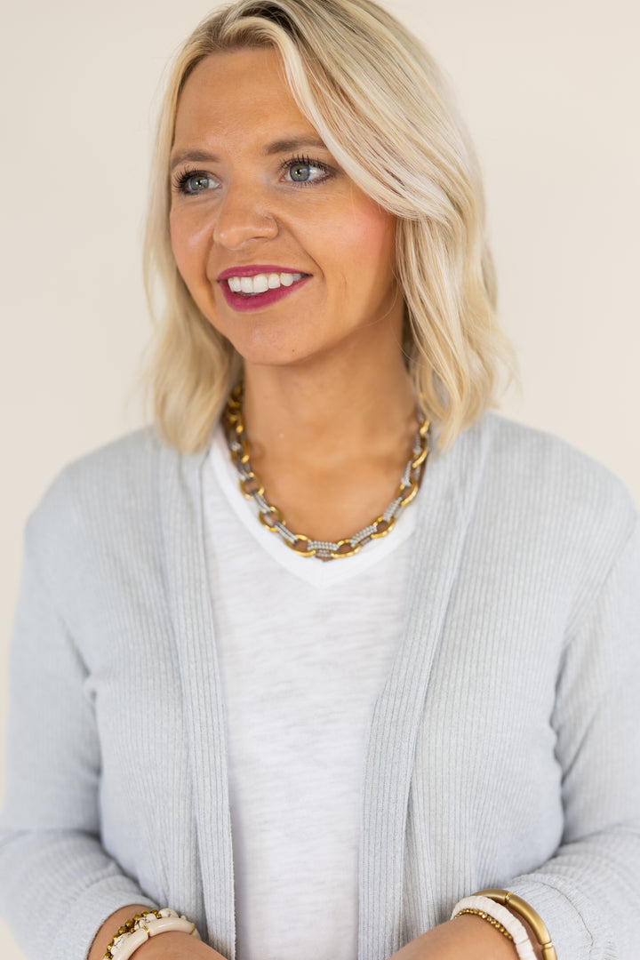 A smiling blonde woman wearing a light gray ribbed cardigan over a white top, accessorized with a chunky gold and silver chain necklace and white bracelets, against a plain beige background.