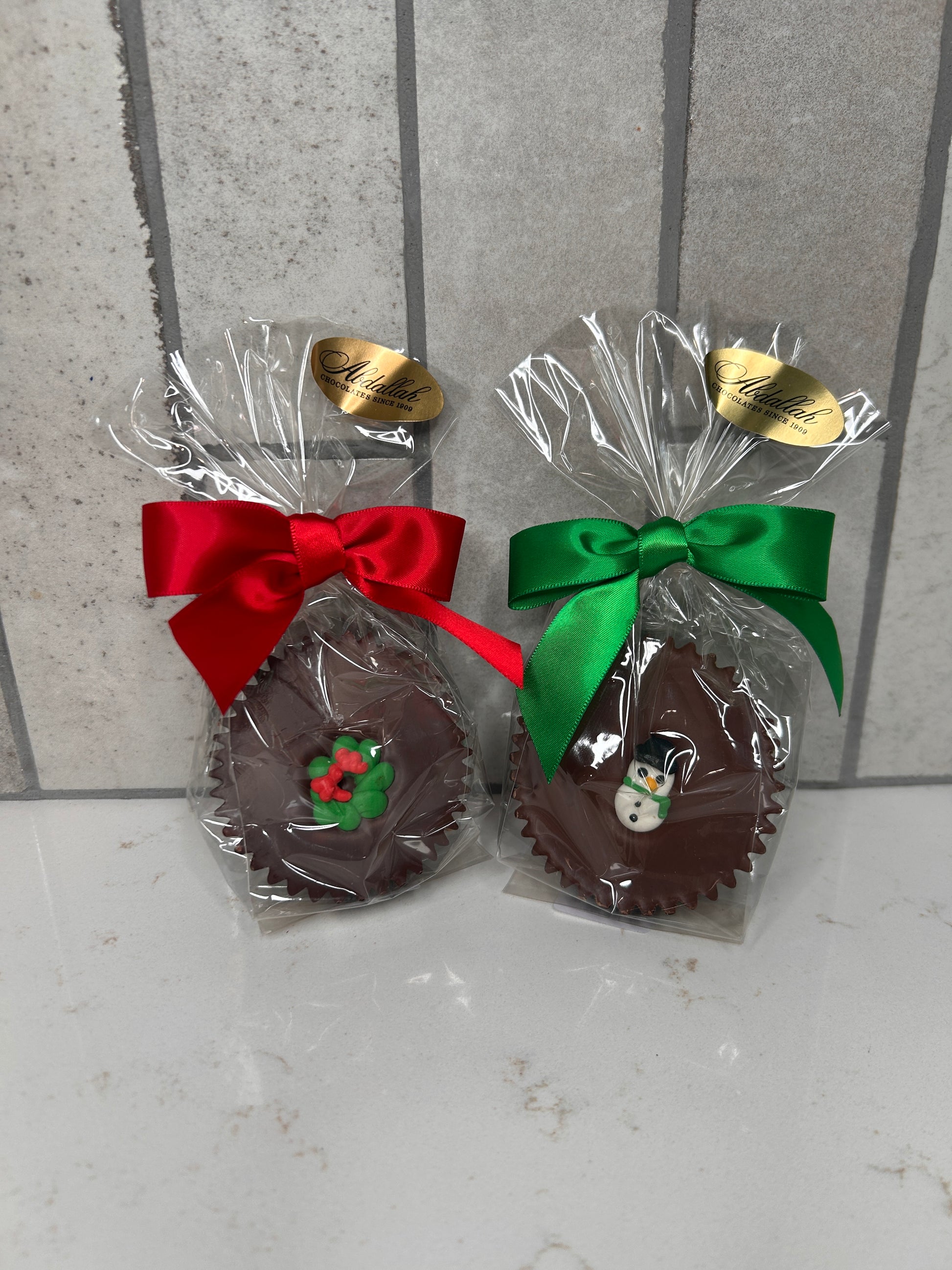 Two individually wrapped chocolate treats, each in clear cellophane with a festive satin bow. The left chocolate features a red bow and a green-and-red holly decoration; the right chocolate has a green bow and a small snowman decoration. Both packages have a gold ‘Chocolates Since 1980’ label and sit on a light tile background.