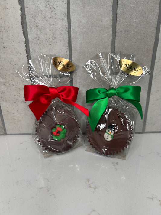 Two individually wrapped chocolate treats, each in clear cellophane with a festive satin bow. The left chocolate features a red bow and a green-and-red holly decoration; the right chocolate has a green bow and a small snowman decoration. Both packages have a gold ‘Chocolates Since 1980’ label and sit on a light tile background.