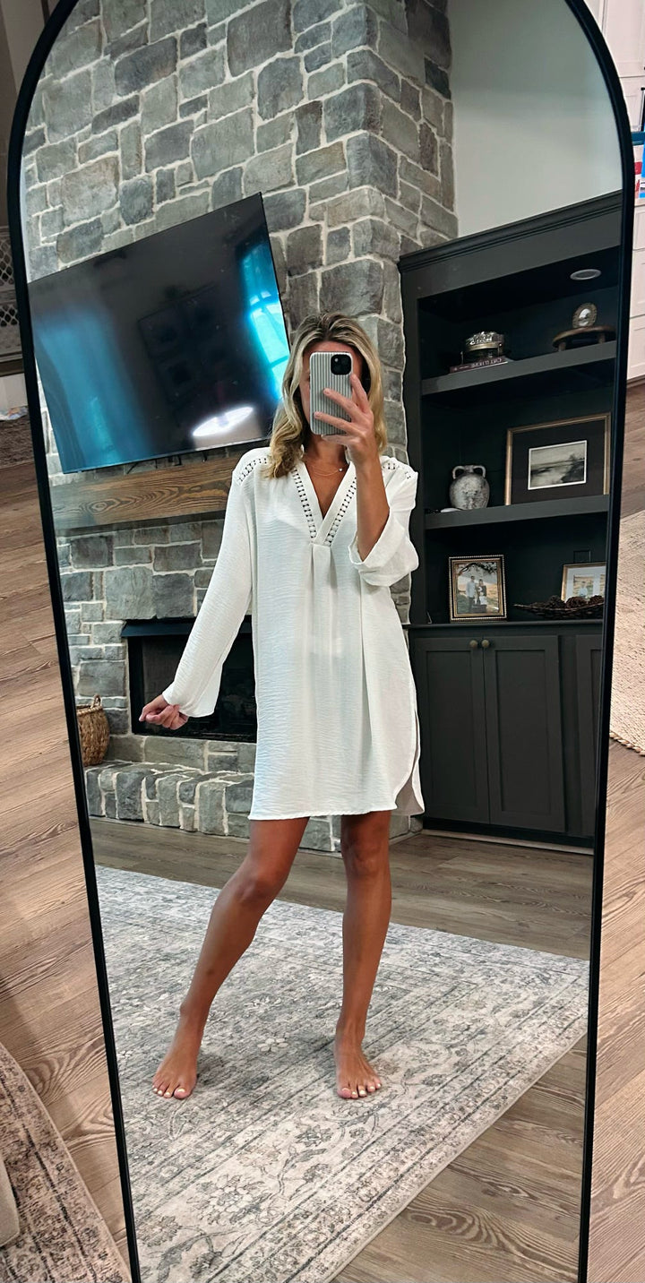 A woman takes a mirror selfie in a living room wearing a white long-sleeve tunic dress with a V-neckline. The background features a gray stone fireplace, a black wooden entertainment cabinet, and a patterned rug.