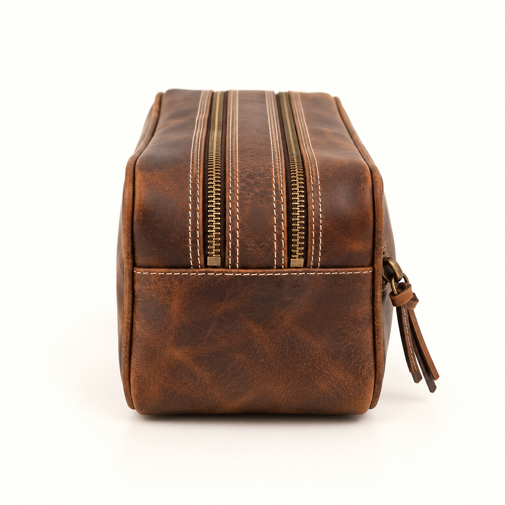 Dusty Dopp Kit men's brown leather toiletry organizer featuring dual antique brass zippers, contrast stitching, distressed leather, and a side carrying loop on a cream background.