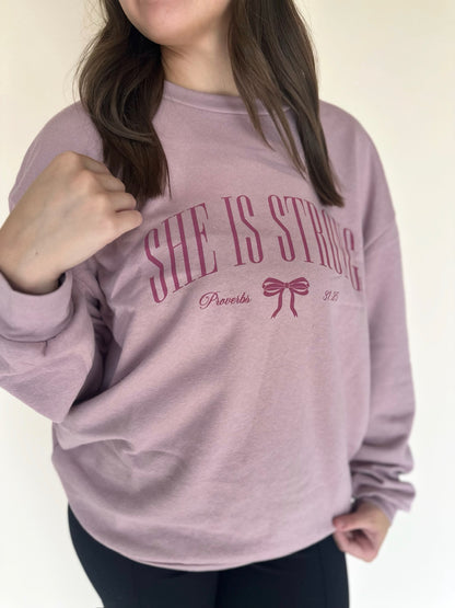 Person wearing a pink sweatshirt with 'SHE IS STRONG' text and a bow graphic.