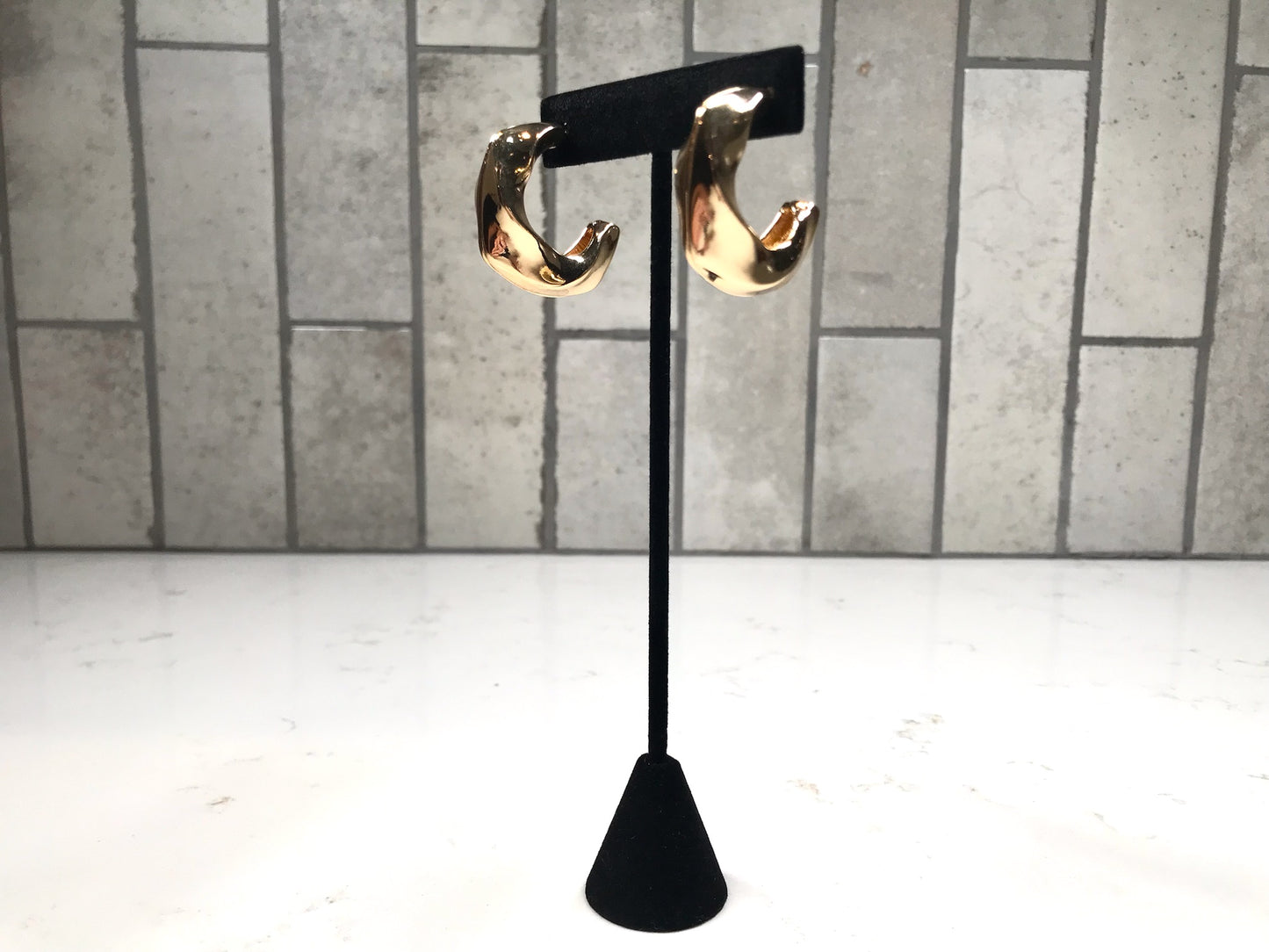 Thick gold sculpted hoop earrings on a black jewelry stand with a neutral tile backdrop.