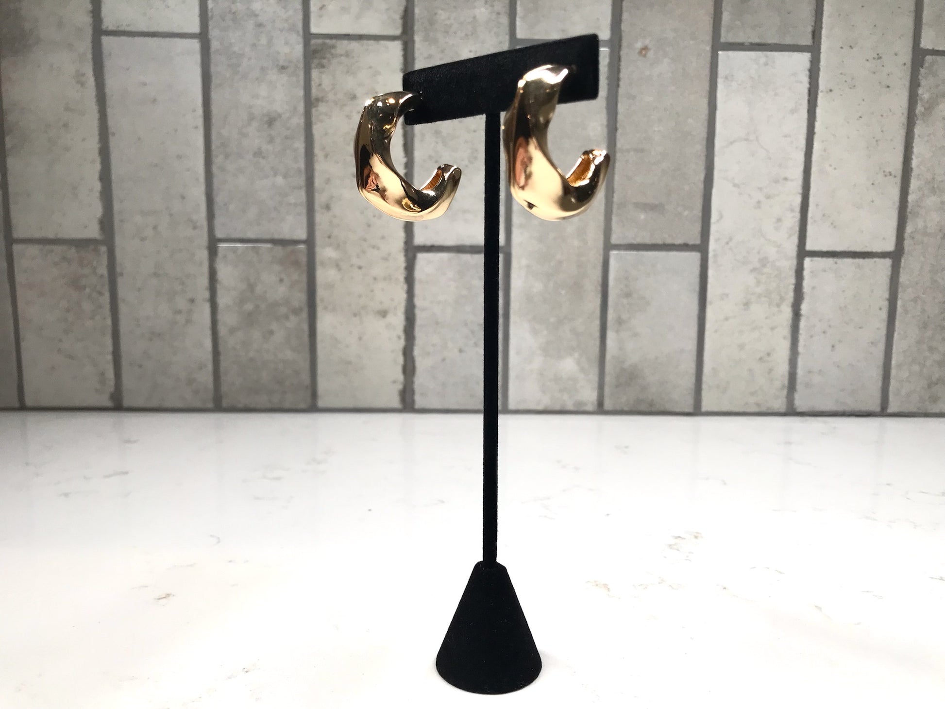 Thick gold sculpted hoop earrings on a black jewelry stand with a neutral tile backdrop.