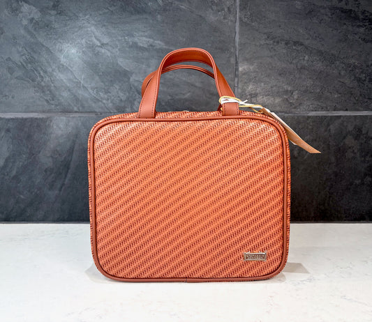Stephanie Johnson Large Briefcase cosmetic travel bag in cognac woven texture with double handles and zip closure, displayed on a countertop with dark tile background.