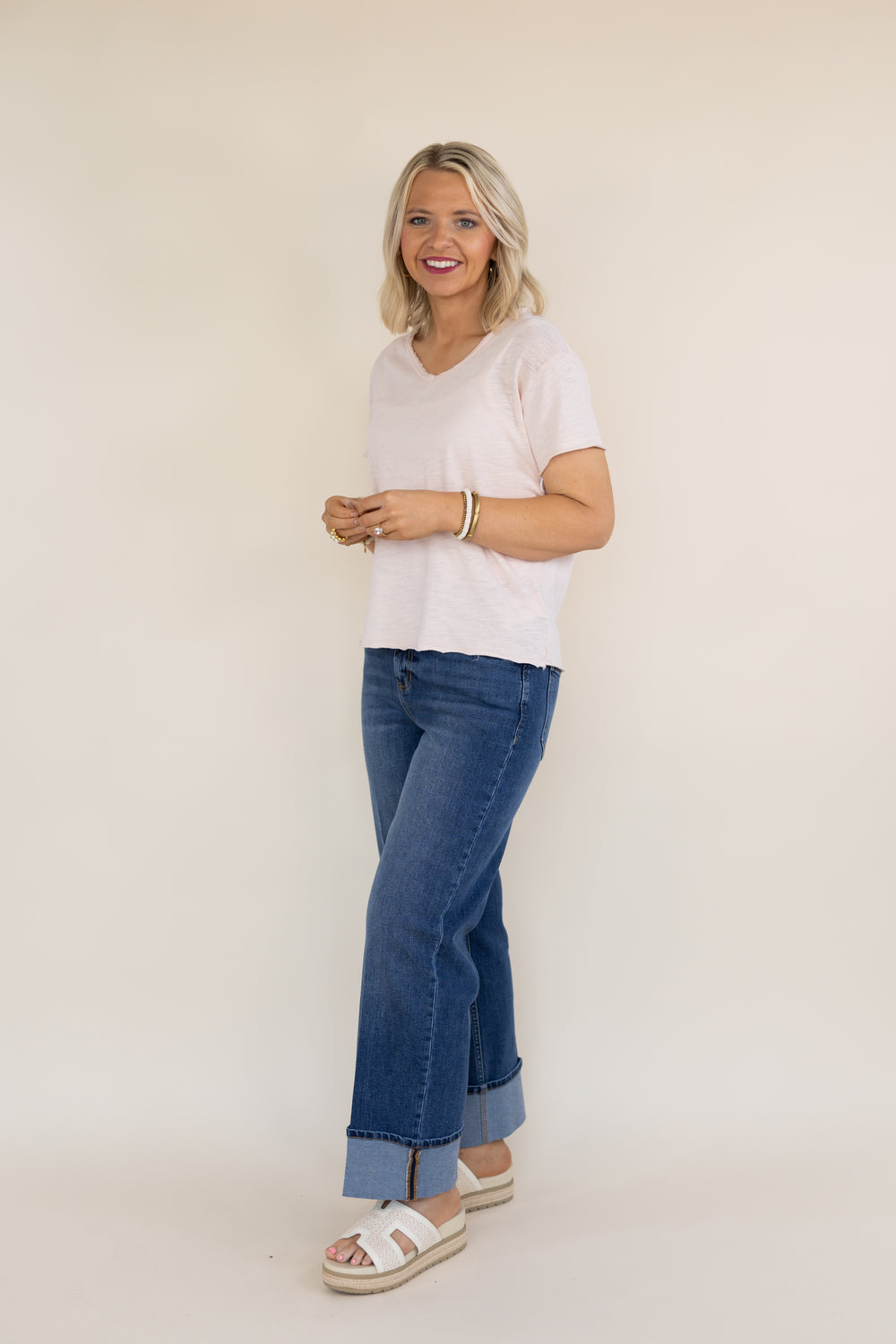 A woman modeling a light pink short-sleeve V-neck t-shirt, blue cuffed jeans, and white slide sandals against a plain beige studio background.