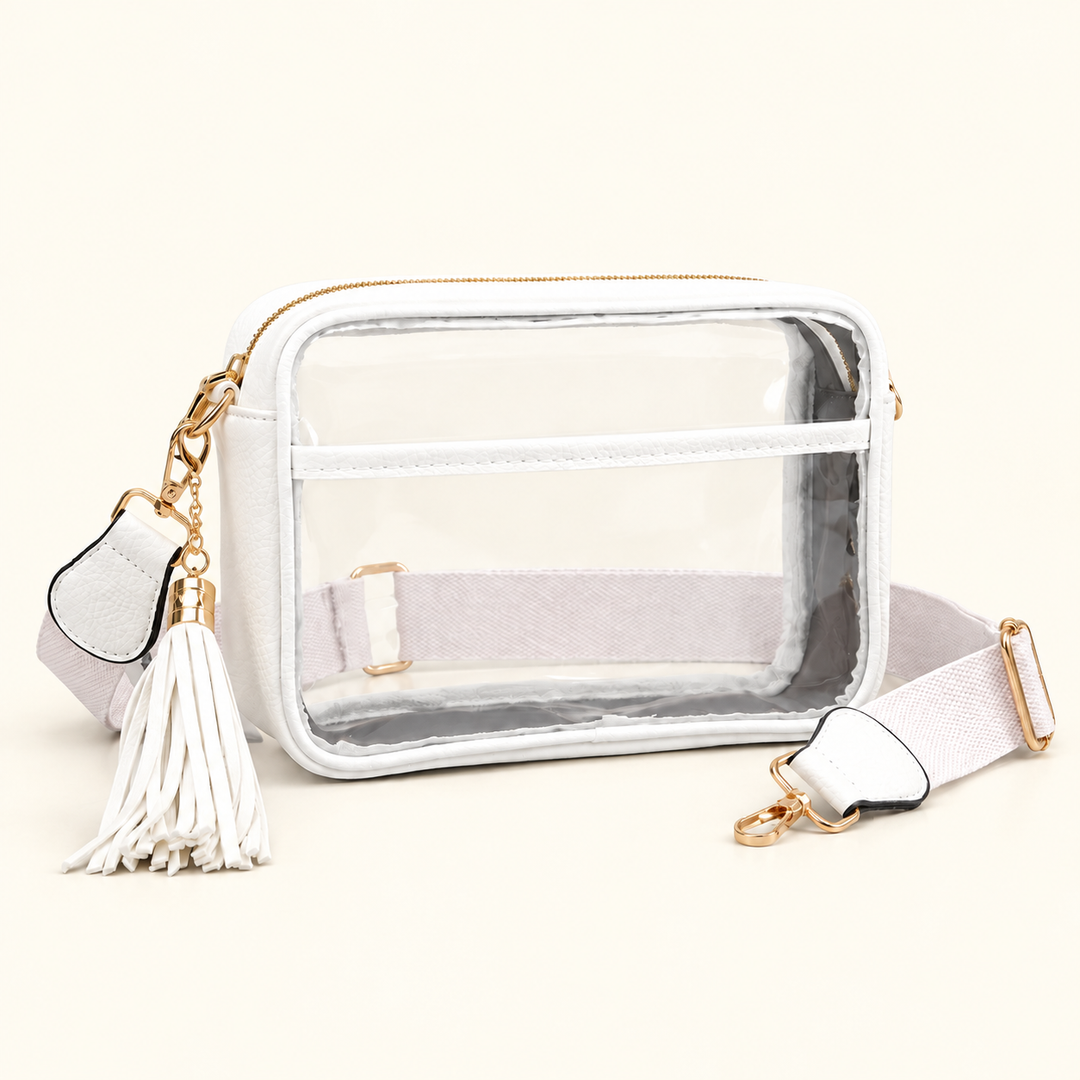 Clear stadium crossbody bag with white trim, tassel charm, pale woven strap, and gold-tone hardware on a warm cream background.