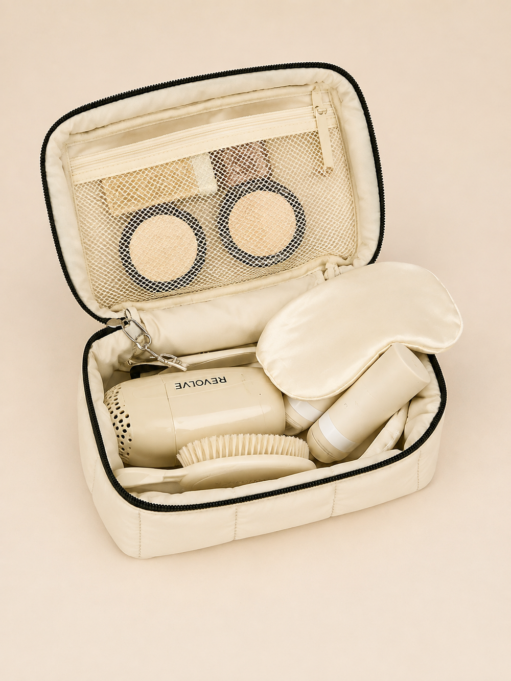 An open cream-colored cosmetic travel case with black trim, containing a hair dryer, brush, mask, and sleep mask, set against a solid cream background.