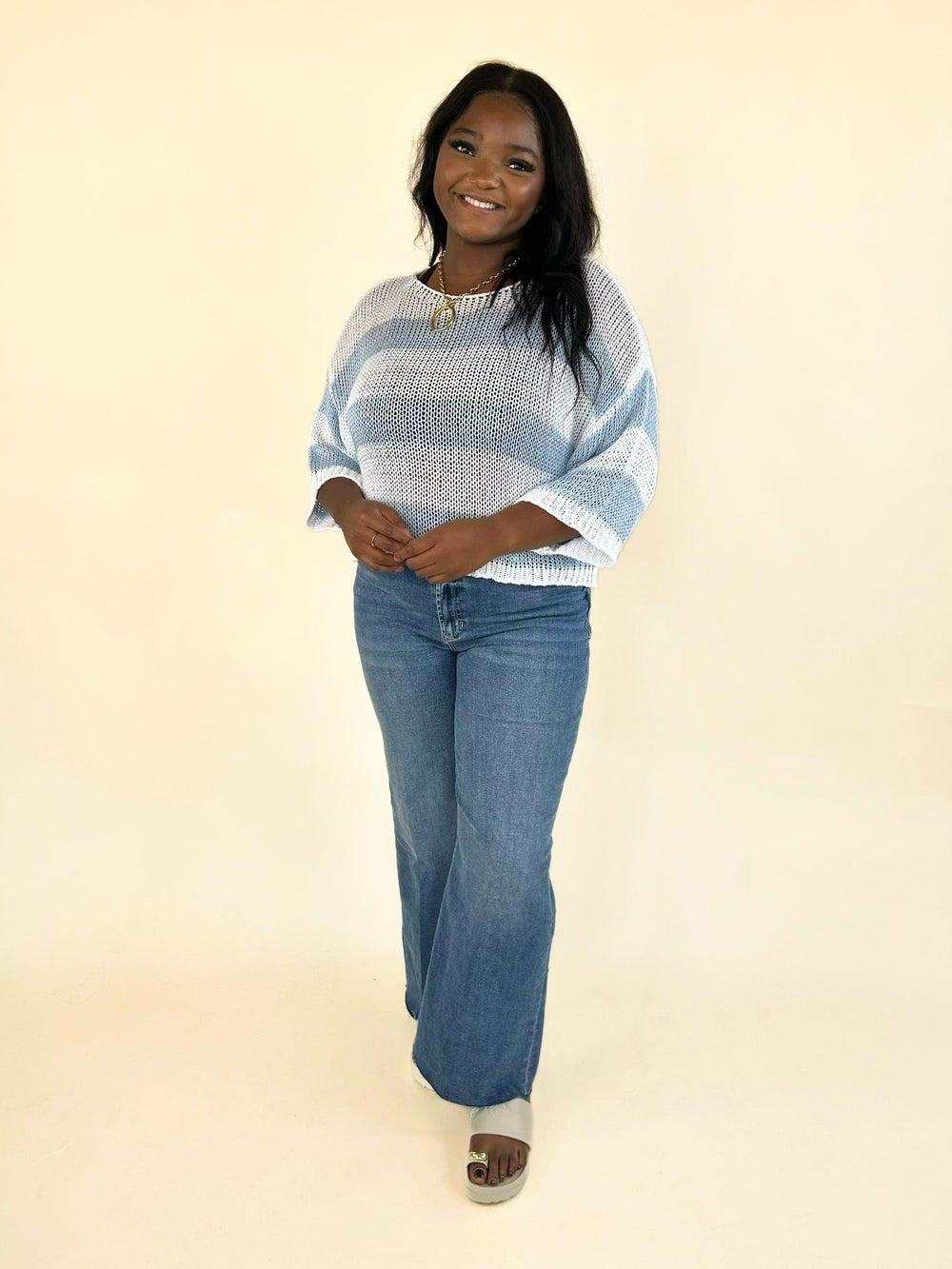 Women's white and light blue striped open-knit sweater with a relaxed silhouette, wide round neckline, airy knit construction, and elbow-length dolman sleeves paired with denim jeans.