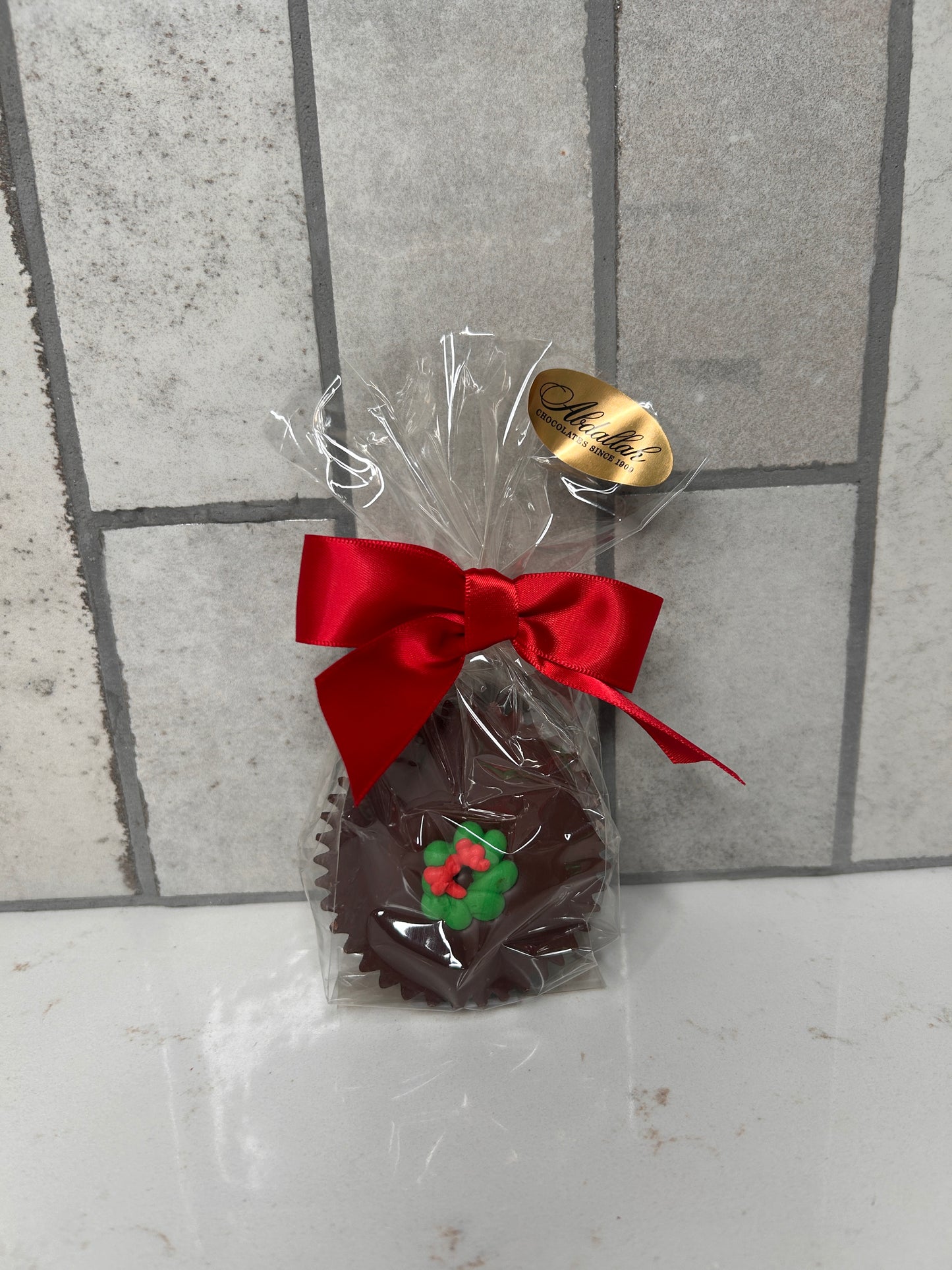 A single chocolate treat wrapped in clear cellophane and finished with a red satin bow. The chocolate is topped with a green-and-red holly decoration. A gold ‘Chocolates Since 1980’ label sits on the packaging, photographed against a light tile background.