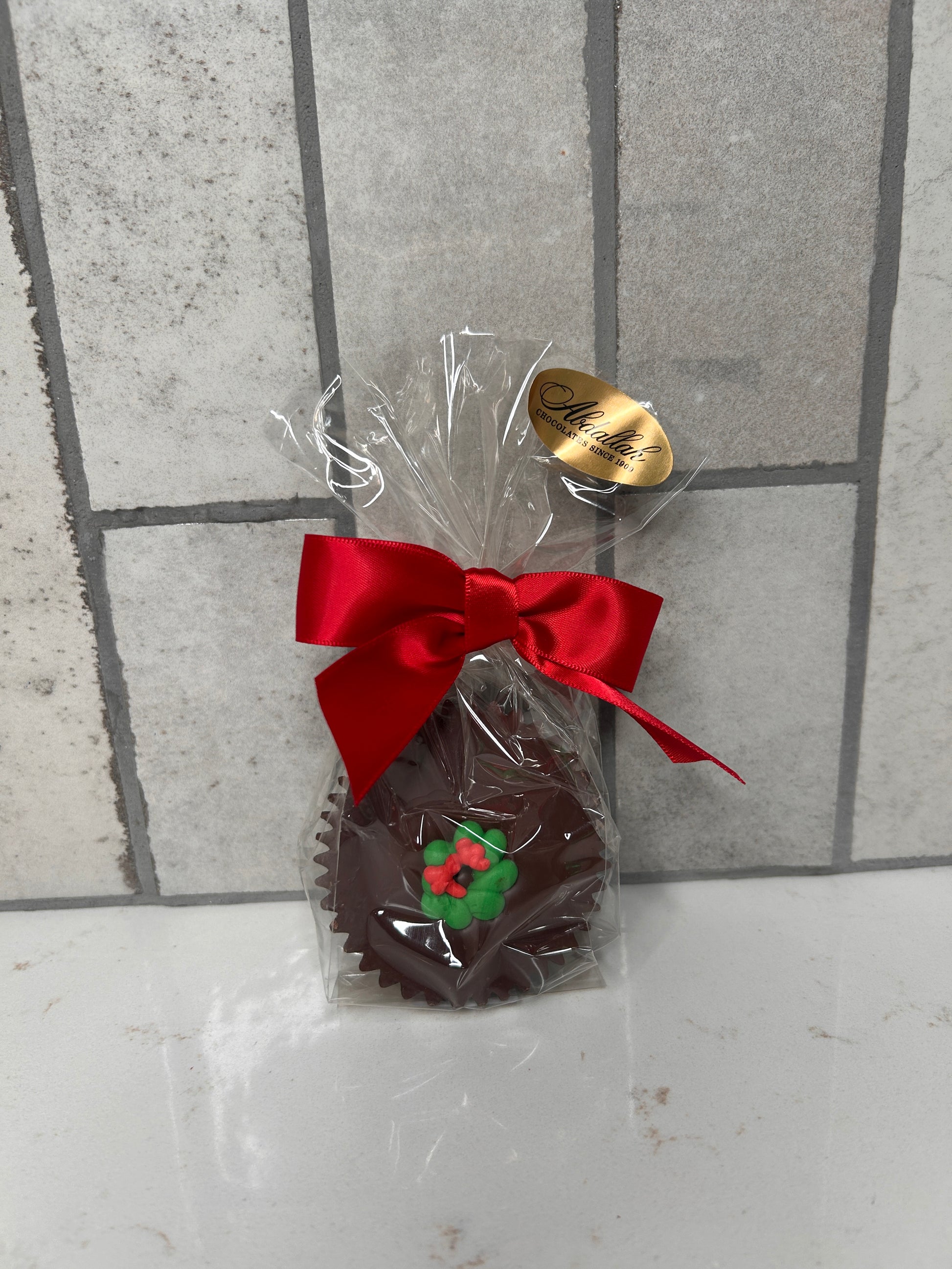 A single chocolate treat wrapped in clear cellophane and finished with a red satin bow. The chocolate is topped with a green-and-red holly decoration. A gold ‘Chocolates Since 1980’ label sits on the packaging, photographed against a light tile background.