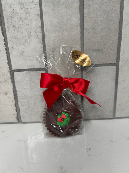 A single chocolate treat wrapped in clear cellophane and finished with a red satin bow. The chocolate is topped with a green-and-red holly decoration. A gold ‘Chocolates Since 1980’ label sits on the packaging, photographed against a light tile background.