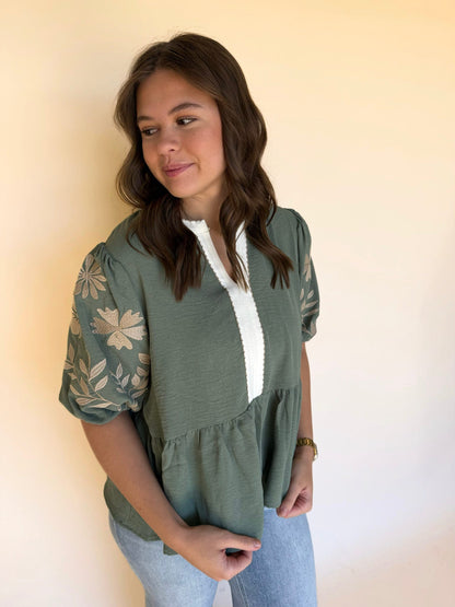Woman wearing a sage green blouse with embroidered sleeves and a white textured neckline, styled with light wash jeans.