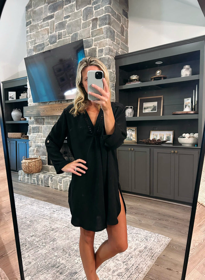 A woman takes a mirror selfie in a living room wearing a black long-sleeve mini dress with a V-neckline and side slits. The background features a stone fireplace, a mounted TV, and dark gray built-in shelves.