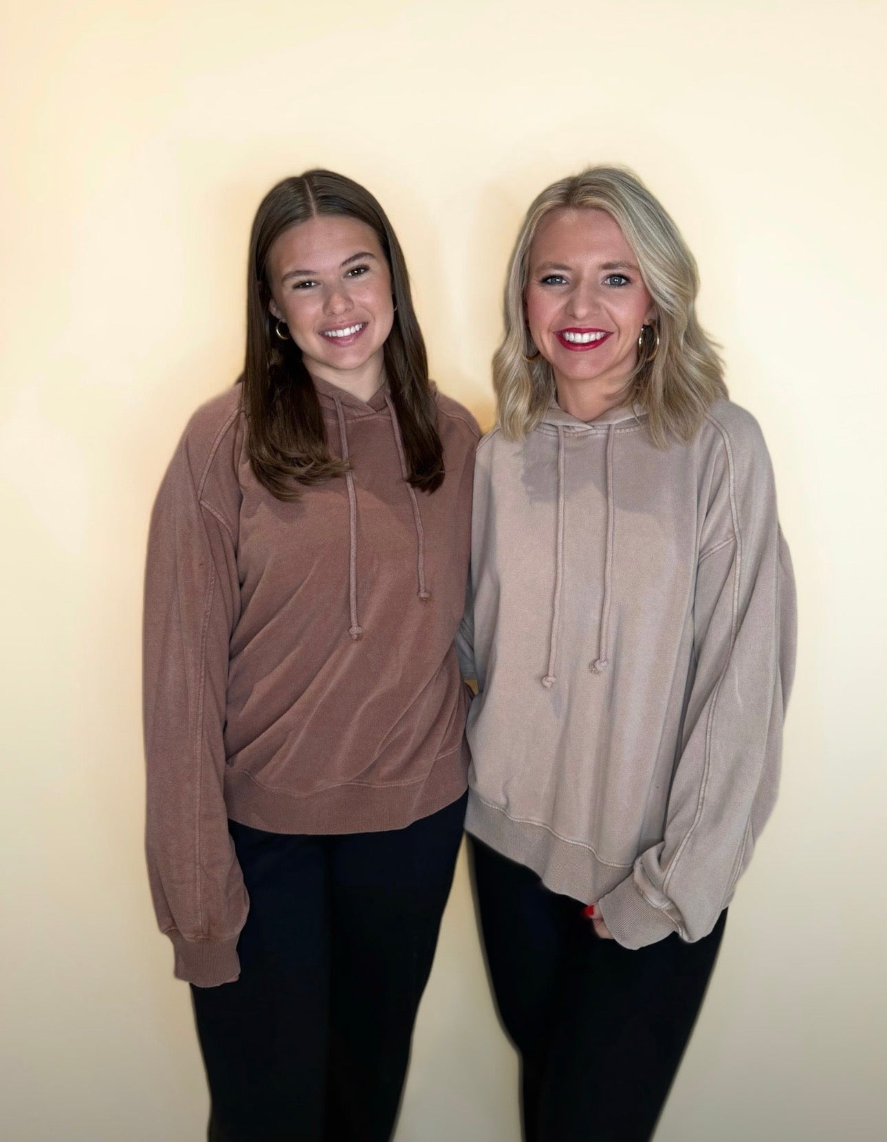 Two women standing side-by-side wearing oversized hoodies. One is in a washed mocha brown hoodie and black pants, and the other in a washed taupe hoodie with black pants. Both are smiling against a neutral cream background.