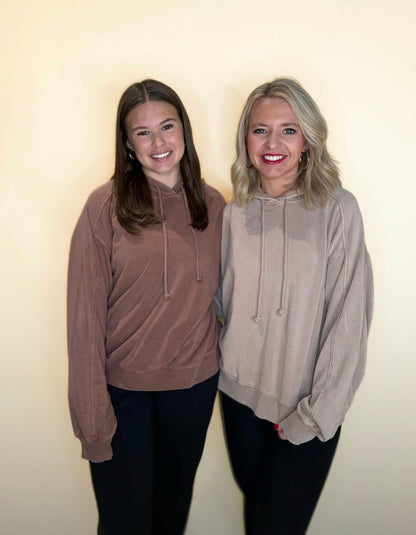 Two women standing side-by-side wearing oversized hoodies. One is in a washed mocha brown hoodie and black pants, and the other in a washed taupe hoodie with black pants. Both are smiling against a neutral cream background.