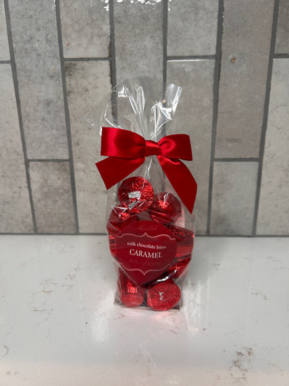 Clear cellophane gift bag filled with red-foil-wrapped caramel milk chocolate bites, tied with a bright red satin bow, sitting on a white countertop with a neutral tile backsplash.