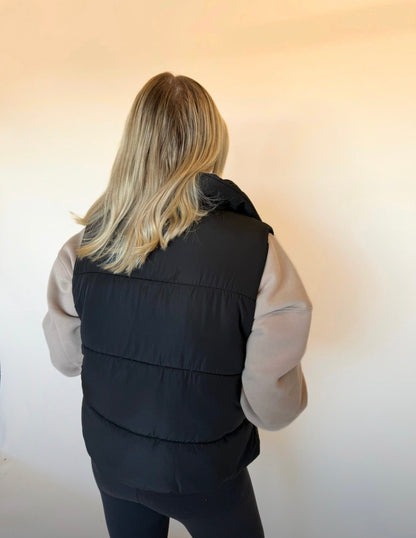 Person wearing a black puffer vest over a light-colored shirt against a plain background
