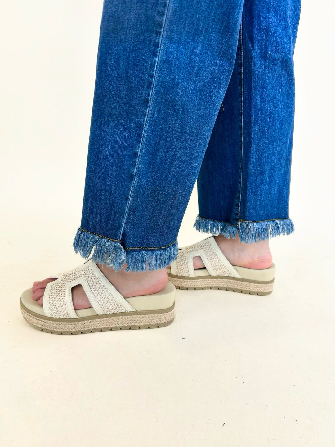 Close-up of the frayed raw hem on medium wash wide leg denim jeans paired with platform sandals.
