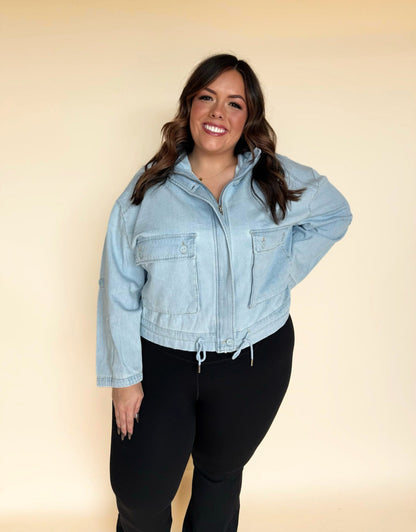 Woman wearing a light-wash denim zip-front jacket with large chest pockets and an adjustable drawstring hem, paired with black leggings against a soft neutral background.