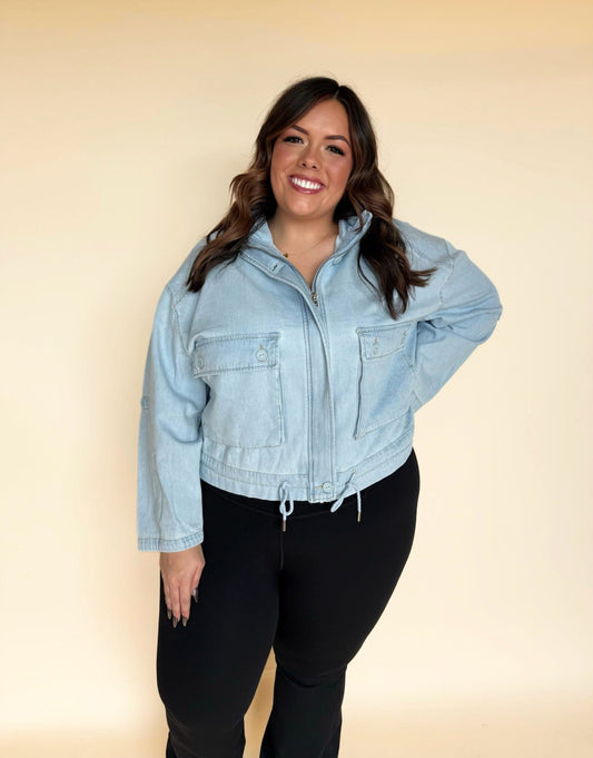 Woman wearing a light-wash denim zip-front jacket with large chest pockets and an adjustable drawstring hem, paired with black leggings against a soft neutral background.