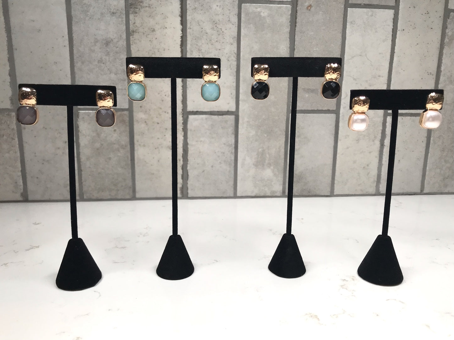 Four pairs of hammered gold stud earrings, each with a different faceted drop stone—gray, light blue, black, and pearl —displayed on black jewelry stands against a light tile background.
