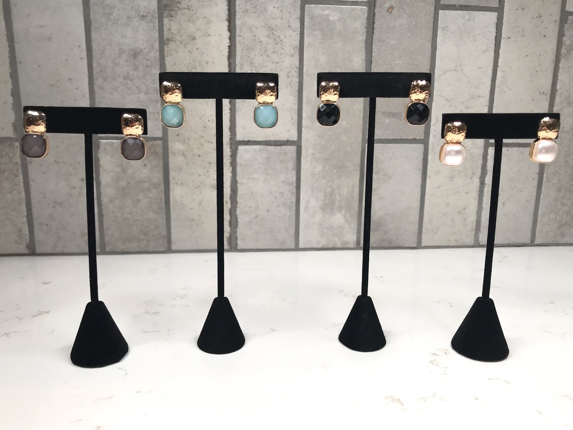 Four pairs of hammered gold stud earrings, each with a different faceted drop stone—gray, light blue, black, and pearl —displayed on black jewelry stands against a light tile background.