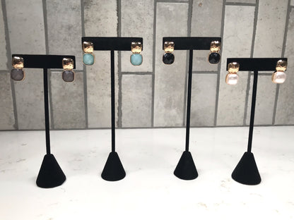 Four pairs of hammered gold stud earrings, each with a different faceted drop stone—gray, light blue, black, and pearl —displayed on black jewelry stands against a light tile background.