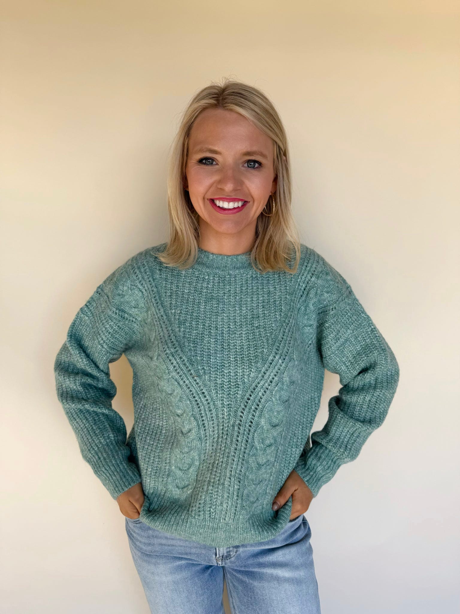 Woman wearing a textured green cable-knit sweater with long sleeves and a crew neckline, paired with light-wash blue jeans against a neutral background.