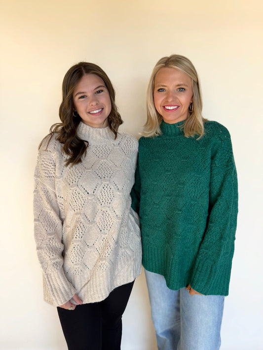 Two women standing together wearing textured knit sweaters; one in a cream mock-neck pullover with a diamond pattern and the other in a green mock-neck sweater with a subtle geometric knit, posed against a neutral background.