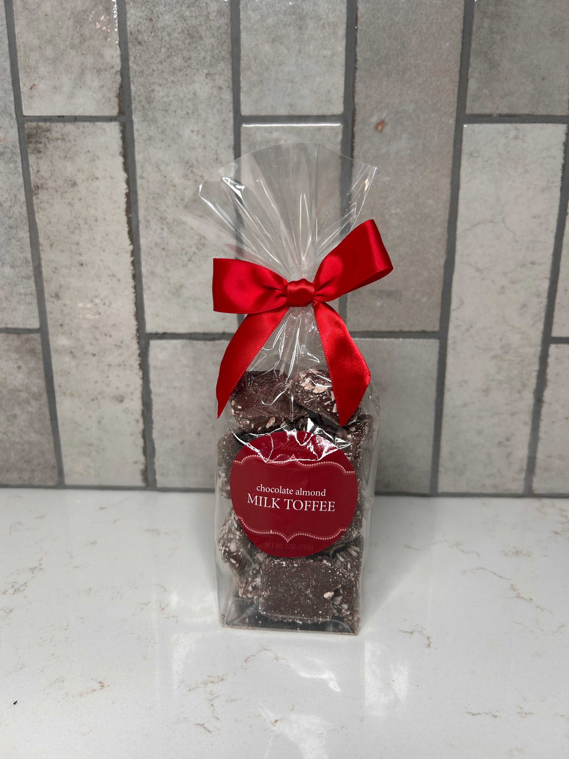 Clear gift bag of chocolate almond milk toffee pieces tied with a red satin bow, featuring a red label, displayed on a light countertop with a neutral tile backsplash.