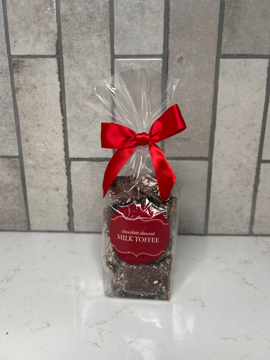 Clear gift bag of chocolate almond milk toffee pieces tied with a red satin bow, featuring a red label, displayed on a light countertop with a neutral tile backsplash.