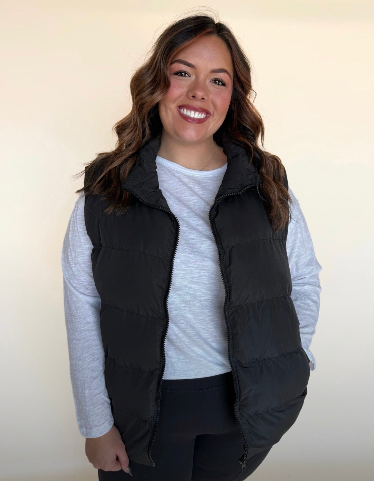Woman wearing a black puffer vest with a white long-sleeve top, smiling toward the camera with one hand relaxed at her side.