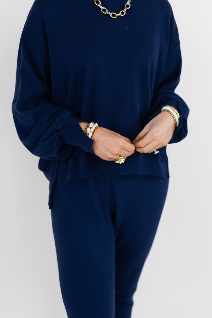 A person wearing a matching navy blue lounge set consisting of a long-sleeve sweatshirt and jogger pants, styled with gold jewelry against a light gray background.
