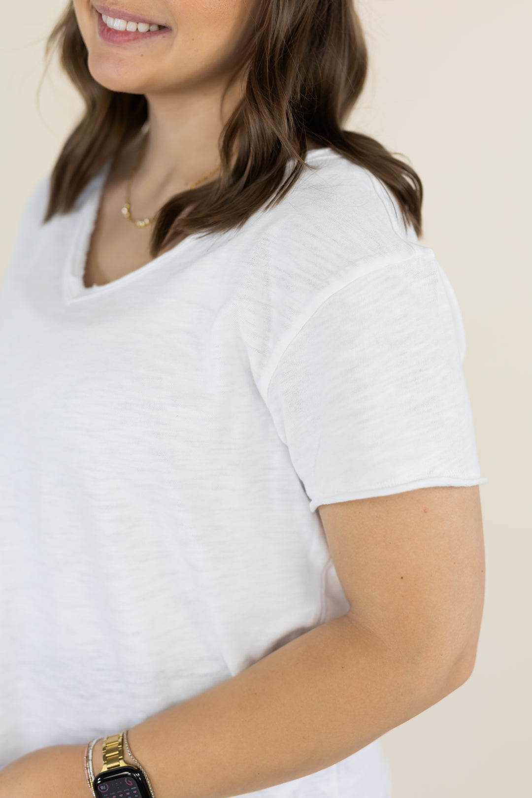 A close-up of a woman wearing a white short-sleeve V-neck t-shirt, showing the fabric texture and fit on her torso against a light beige background.