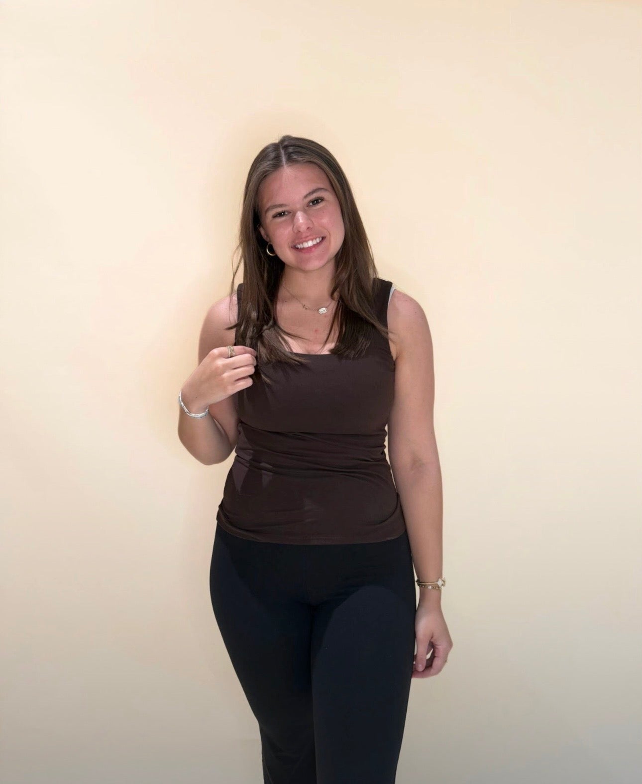 Young woman wearing a fitted brown tank top with black leggings, smiling against a neutral light background.