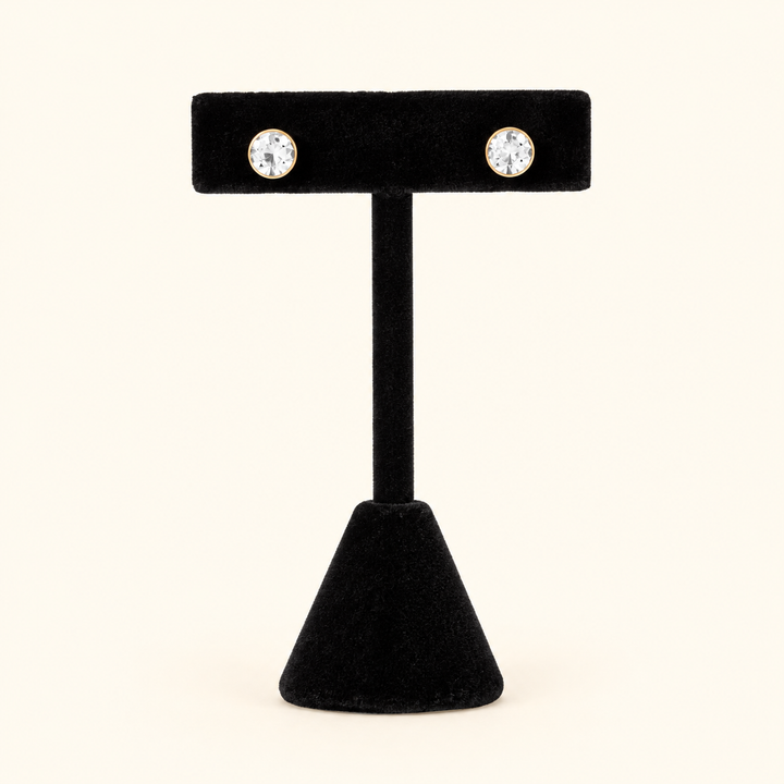 6mm round clear crystal stud earrings with gold-tone settings displayed on a black velvet T-bar jewelry stand against a cream background.