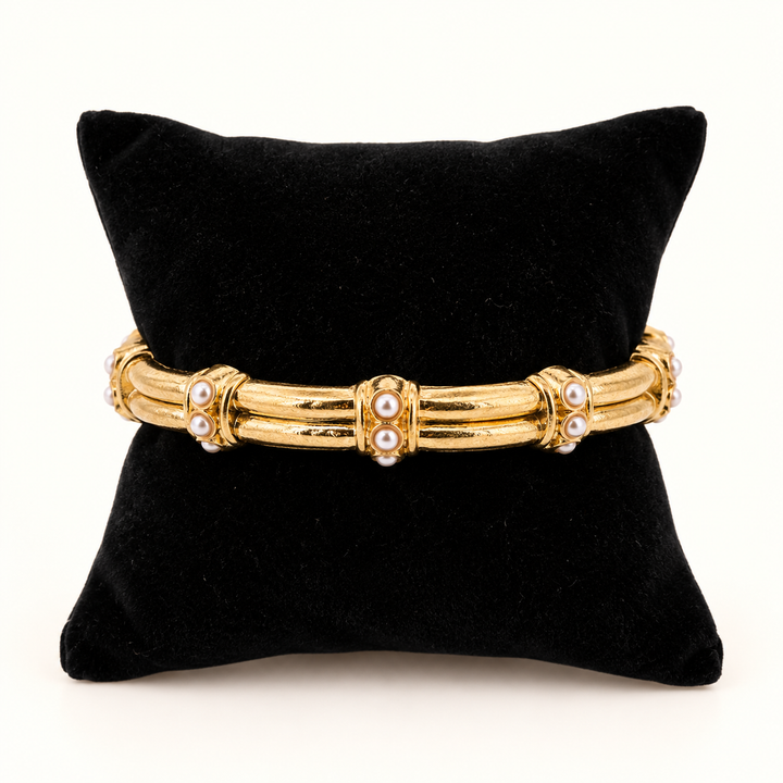 Gianna gold-tone artisan stretch bangle bracelet with white pearl accents and textured gold bars displayed around a black velvet jewelry pillow against a cream background.