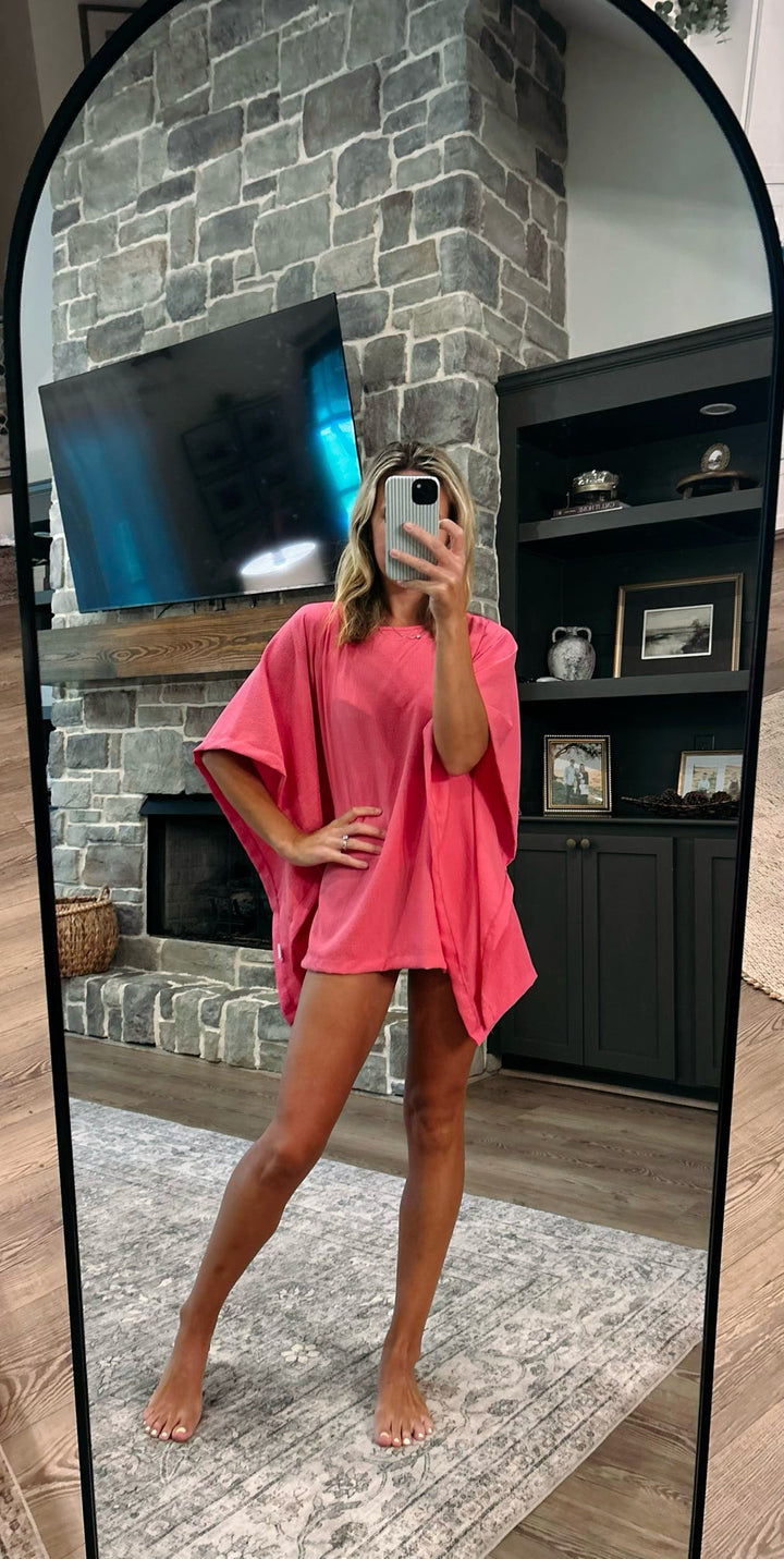 A woman takes a mirror selfie in a living room wearing a bright pink short-sleeve tunic or poncho-style cover-up with a loose, oversized fit and short legs.