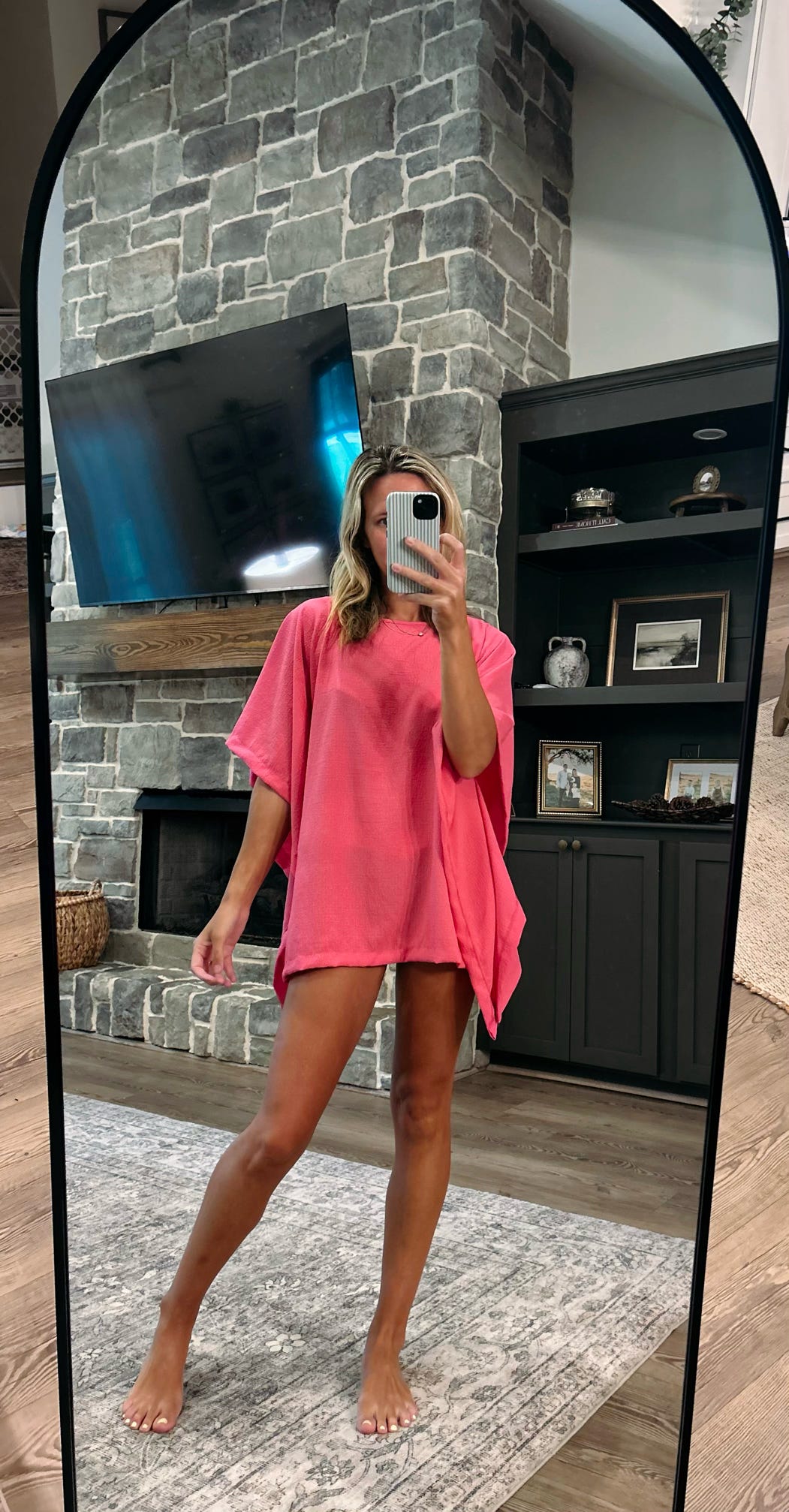 A woman takes a mirror selfie in a living room wearing a bright pink short-sleeve tunic or poncho-style top. The room features a stone fireplace, a mounted TV, and a dark wood cabinet with framed pictures.