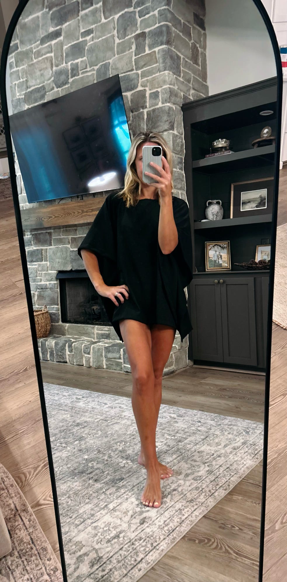 A woman takes a mirror selfie in a living room wearing a loose black short-sleeve tunic dress. The background features a stone fireplace, a black entertainment cabinet, and a gray patterned rug.
