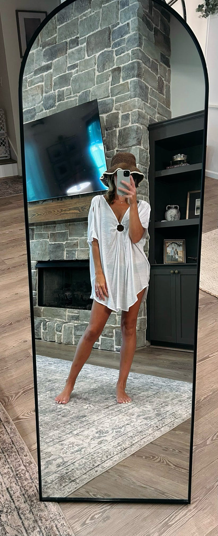 A woman takes a mirror selfie wearing a white short-sleeve tunic dress and a brown wide-brim hat in a living room with a stone fireplace and wood floors.