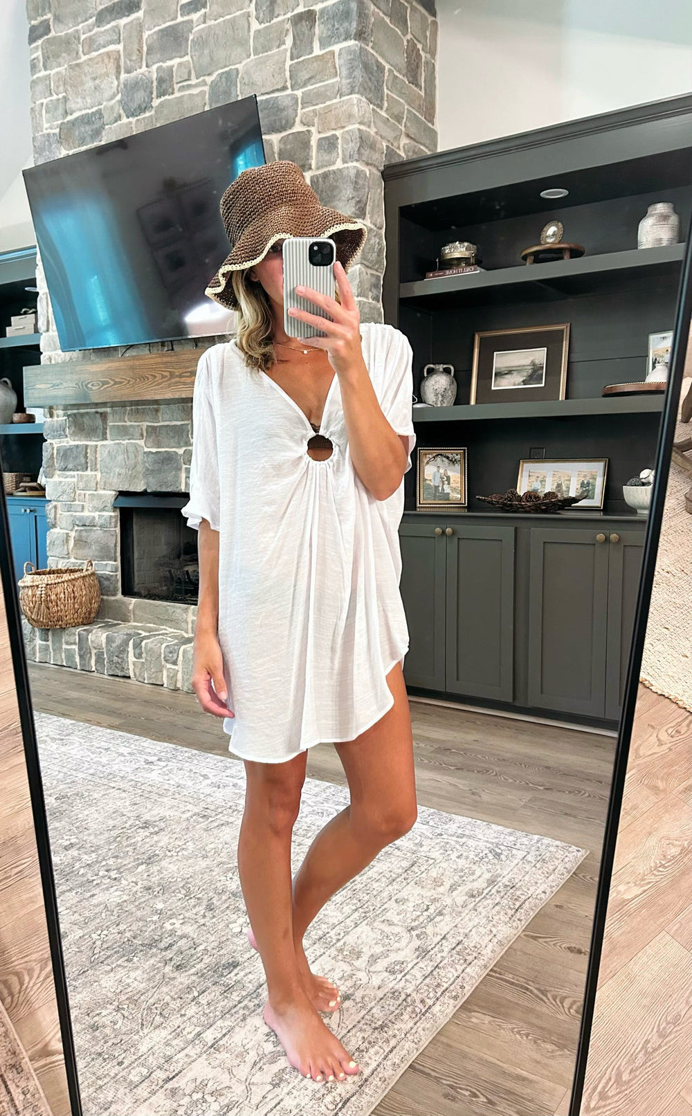 A woman takes a mirror selfie in a cozy living room, wearing a white short-sleeve V-neck tunic dress with a gathered front, a brown wide-brim straw hat, and white sunglasses.